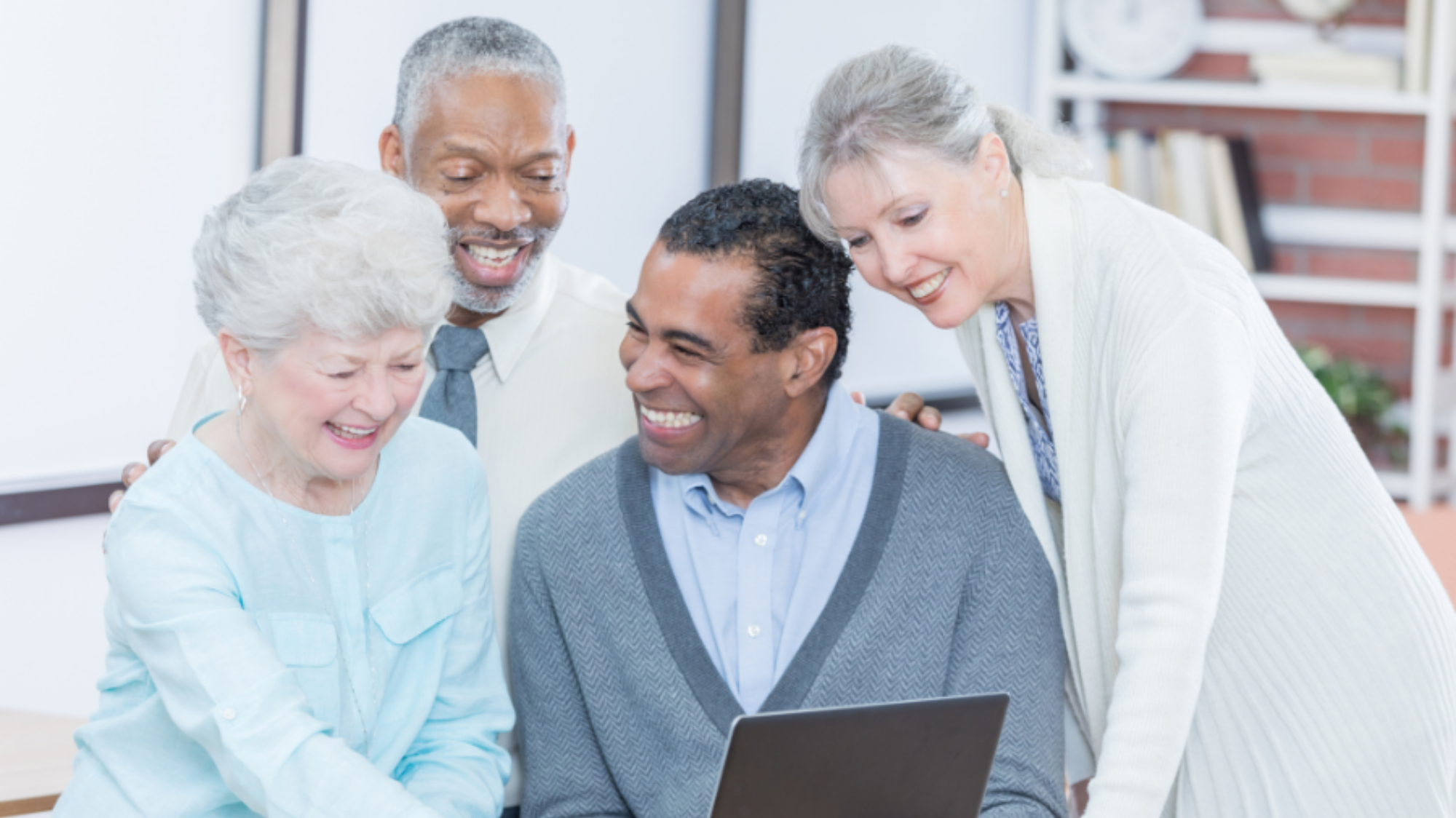 Four older adults gathered around a laptop, wearing expressions of happiness.